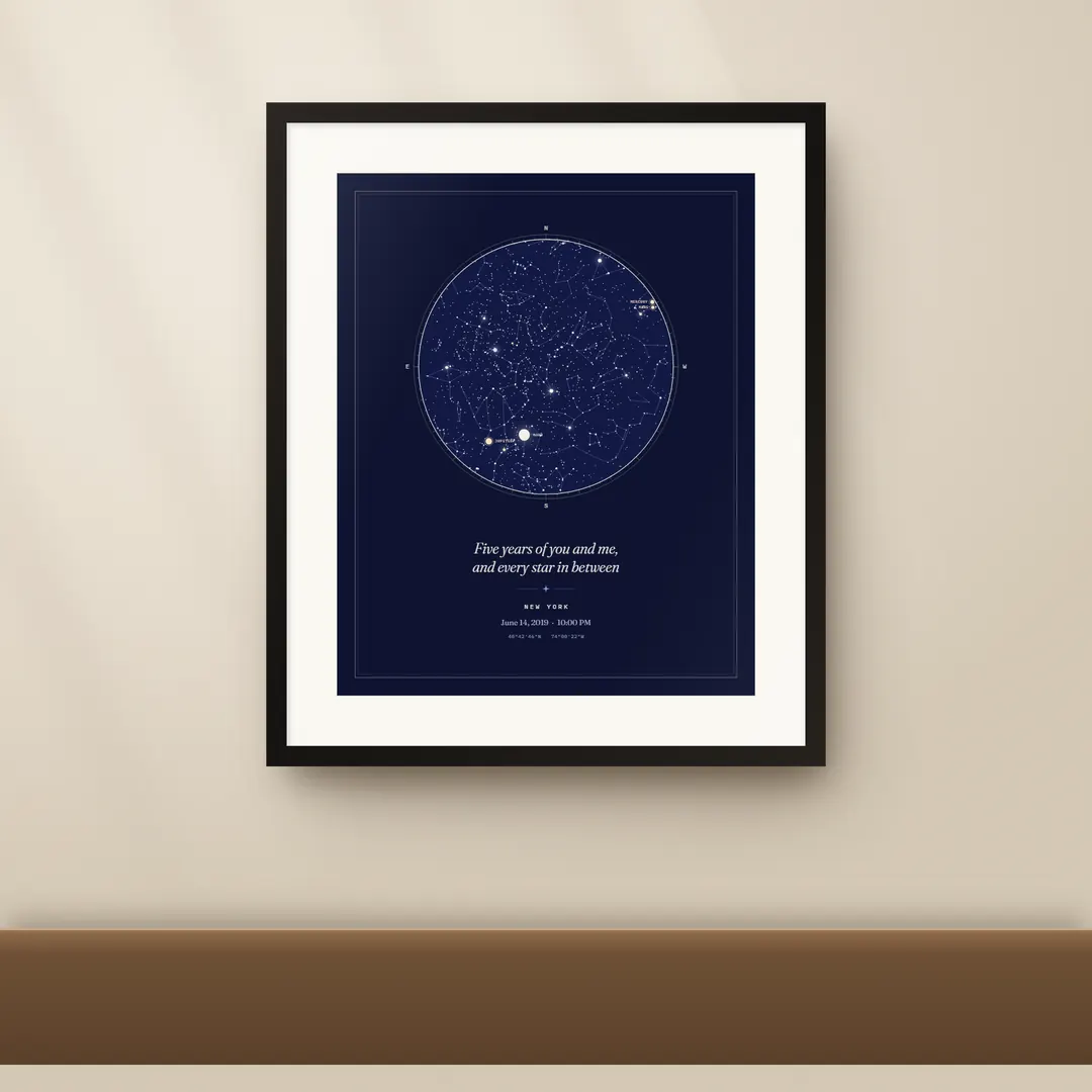 A Your Night Sky print in a black frame with a white mat on a warm wall: a round star map in dark blue, a line of text and the place and date under it.