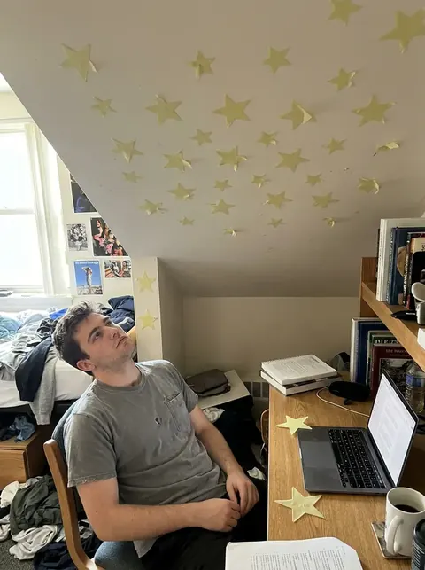 Render of a young man in a dorm room looking up, unimpressed, at faded yellow plastic glow stars on the ceiling, two fallen on his desk.